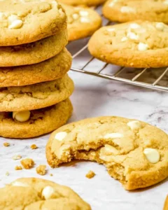 White chocolate chip cookies stacked up, some on a wore rack and one with a big bite out of it.