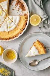 A slice of lemon pie on a plate, which has been taken from a pie dish with the remaining pie still in it.