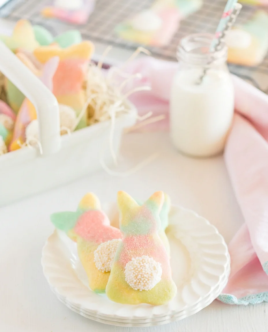 Two pastel bunny-shaped cookies with sugar pearl tails on a white scalloped plate.