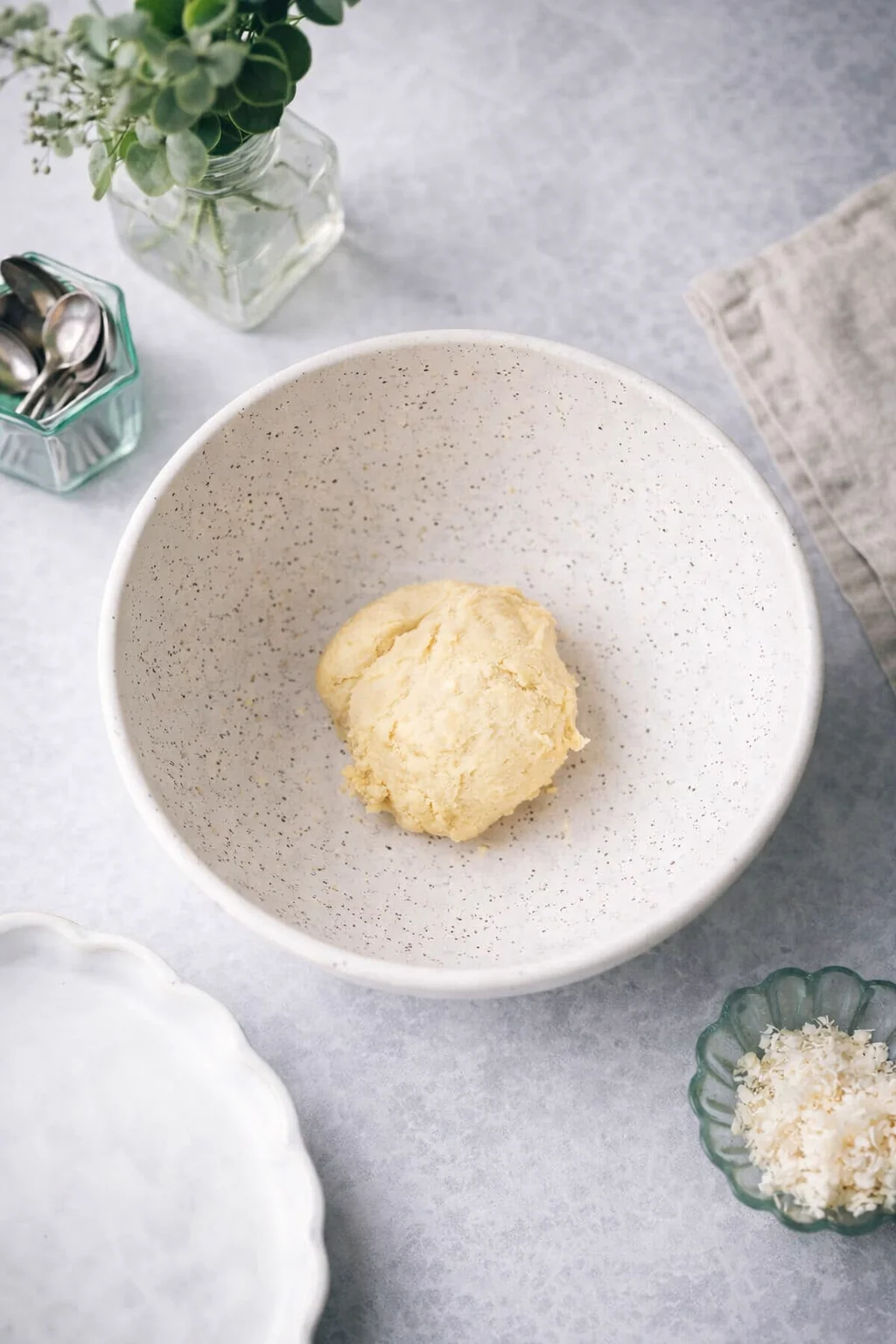 A bowl with a scoop of pale yellow dough or batter on a light gray countertop, surrounded by kitchen tools and decor.