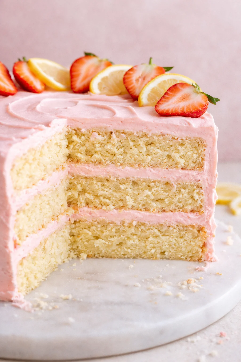 Close-up of a three-layer vanilla cake with pink frosting and strawberries and lemon slices garnish.