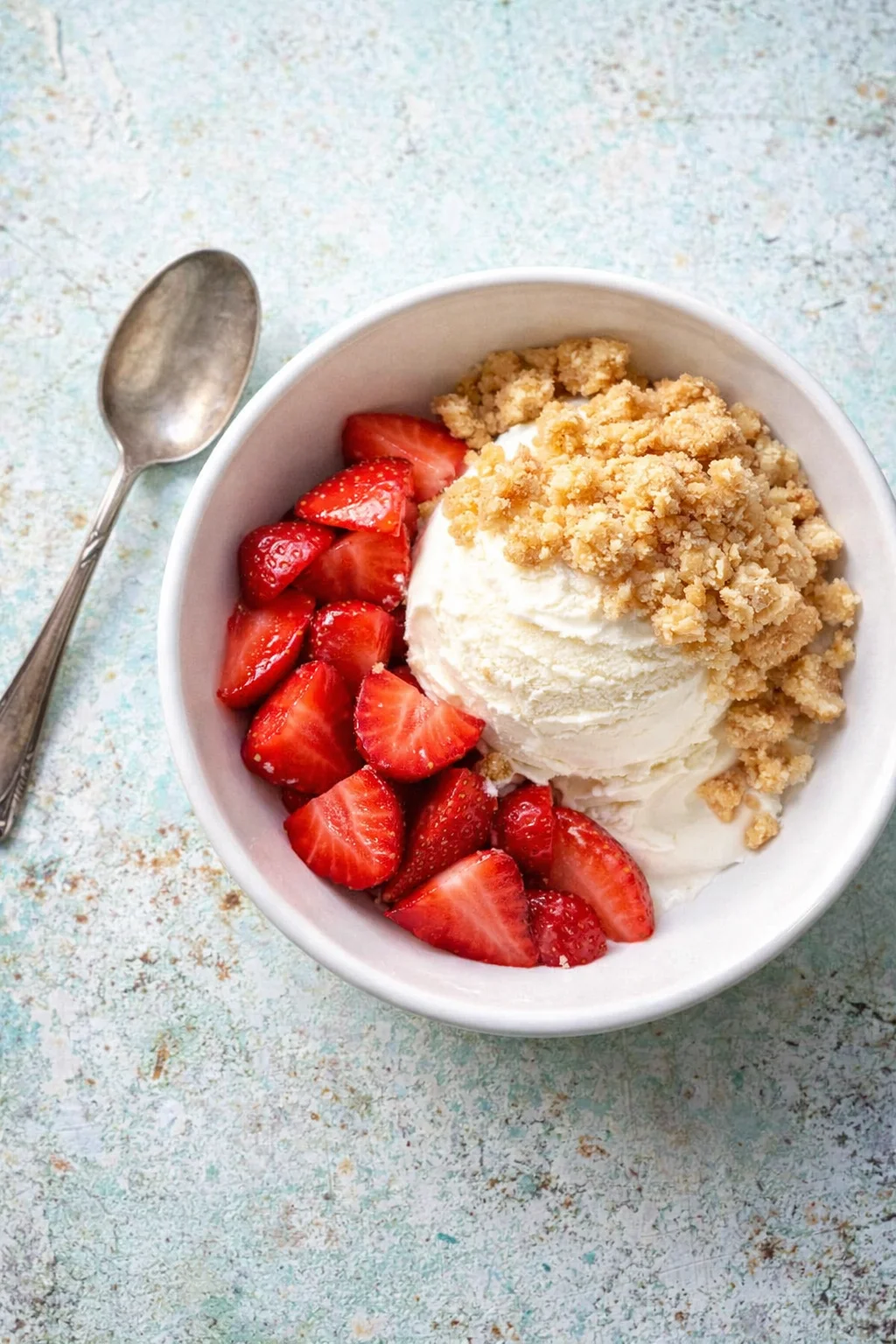 white bowl with vanilla ice cream, sliced strawberries, and crumbly topping on a blue rustic background