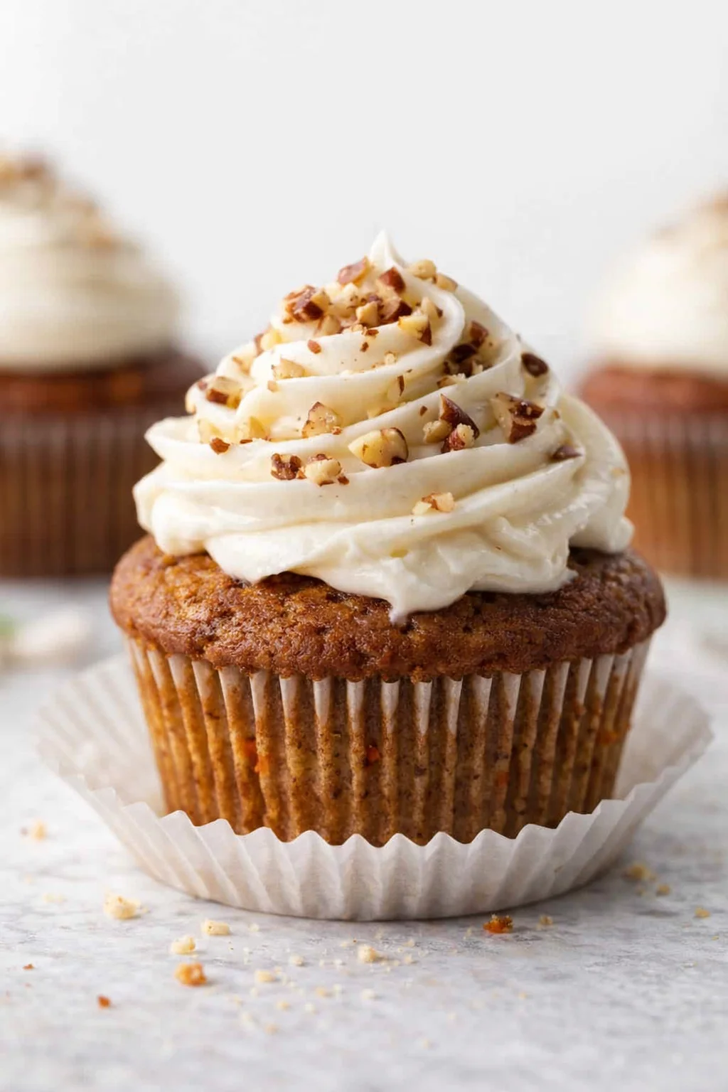 A moist carrot cupcake with white cream cheese frosting and chopped nuts on top.