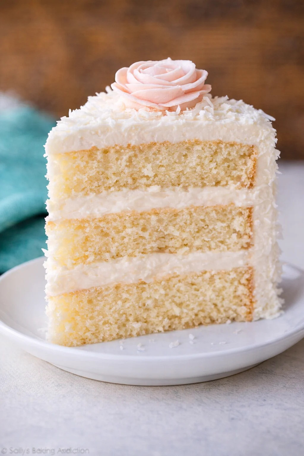 coconut-cake-6.jpg slice of vanilla cake with white frosting and a pink frosting rose on a plate
