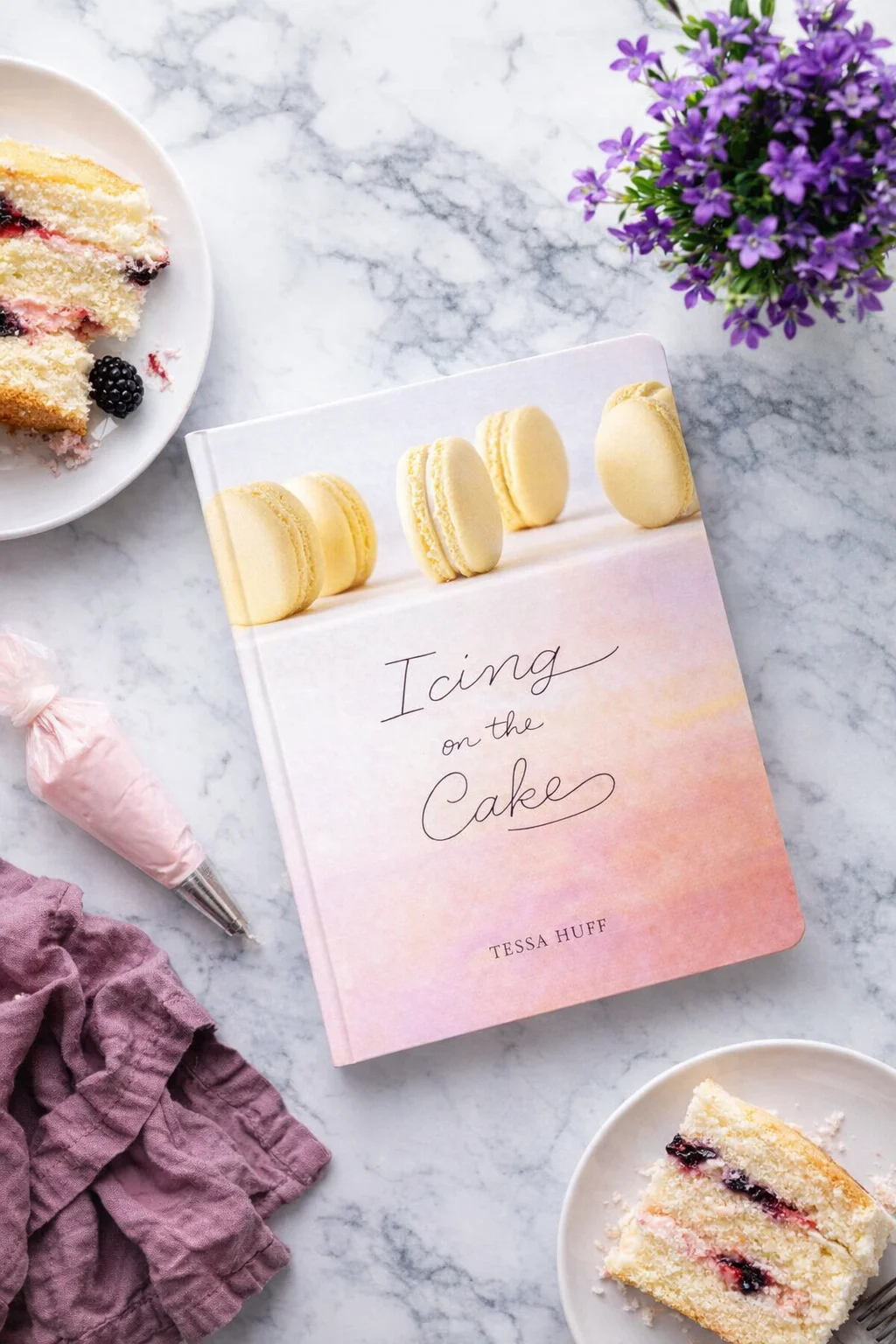 icing-on-the-cake-cookbook.jpg flatlay of pastel cookbook titled icing on the cake with yellow macarons and purple flowers
