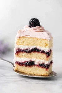 slice of vanilla cake with pink frosting, white cream layers, and berry jam, topped with a blackberry.