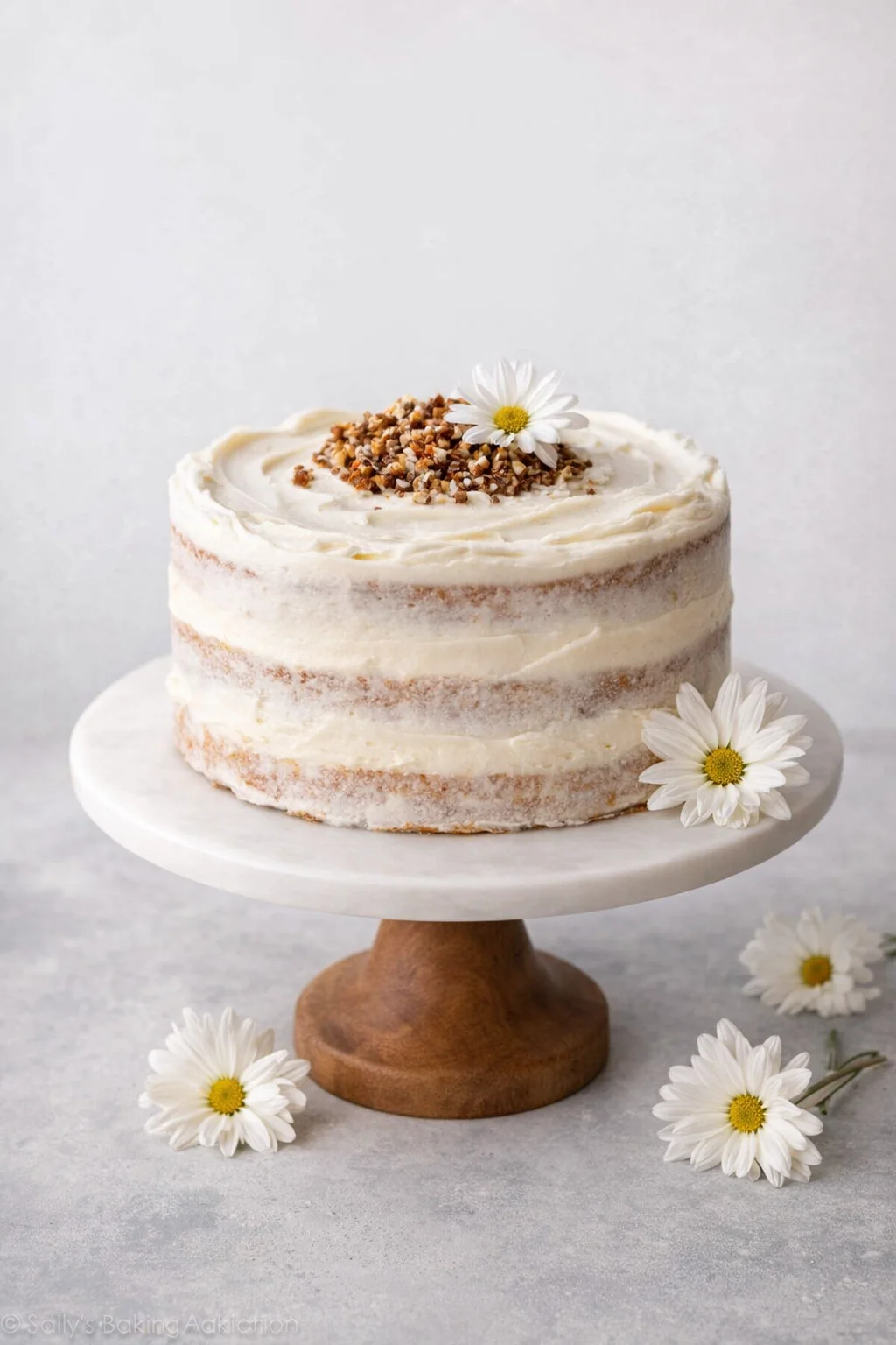 Two-layer naked vanilla cake on a white cake stand, topped with chopped nuts and white flowers.
