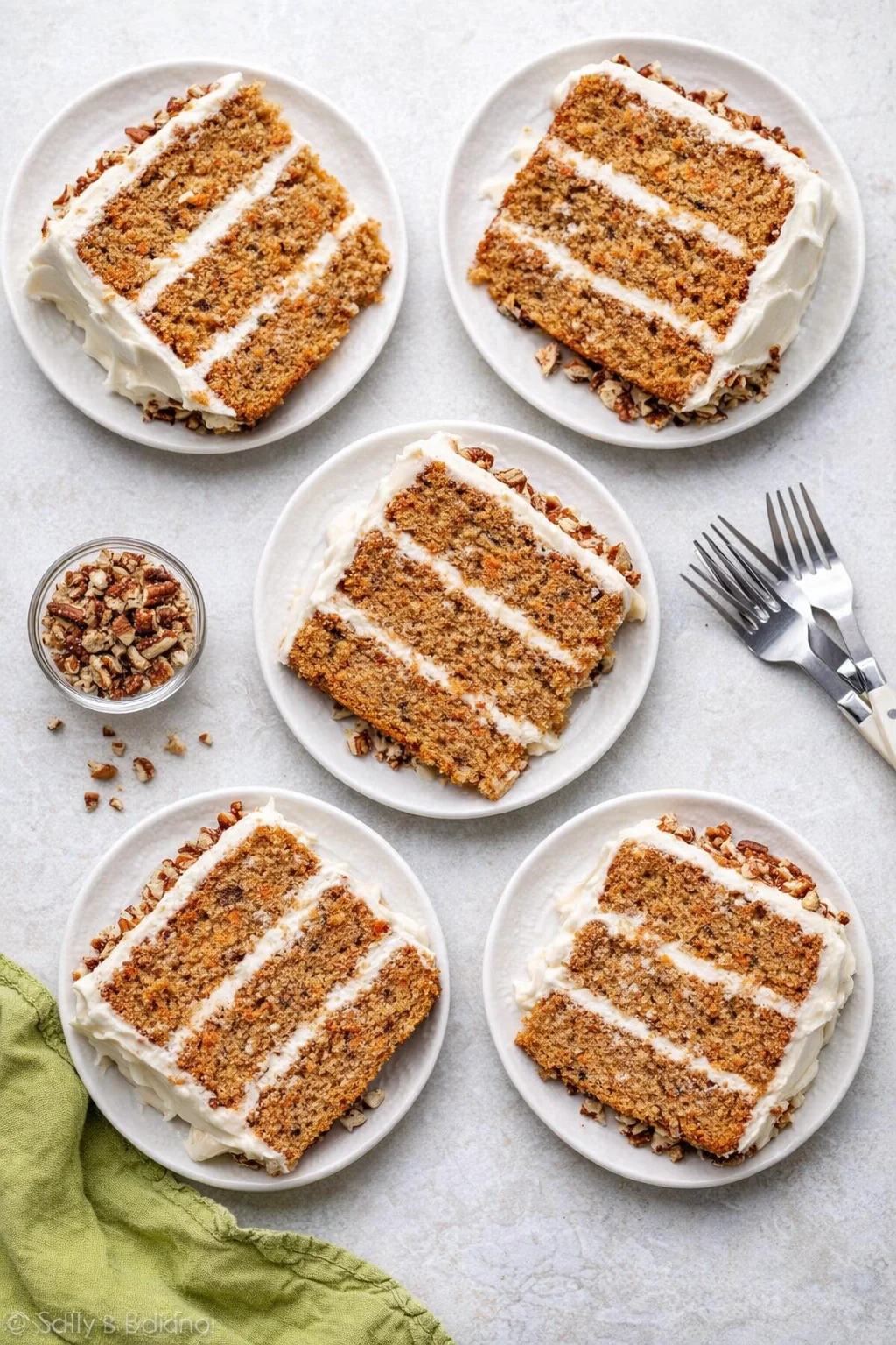 Sliced carrot cake with cream cheese frosting on white plates, nuts scattered, green napkin nearby.