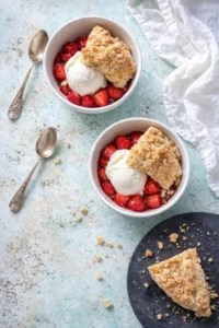 Two bowls of strawberry dessert with vanilla ice cream and crumble on a turquoise surface.