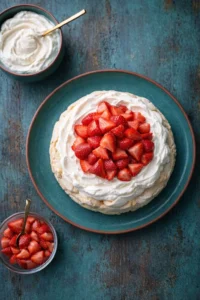 Top-down view of strawberry pavlova on a teal plate, topped with whipped cream and fresh strawberries.