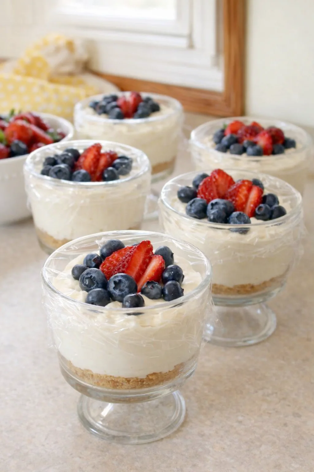 Plastic-wrapped cheesecake cups chilling in refrigerator, some shown topped with fresh berries.