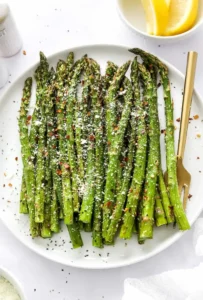 Air fryer asparagus on a round white plate topped with grated cheese and red pepper flakes with a gold fork on the plate next to it and some lemon slices and salt and pepper behind it.