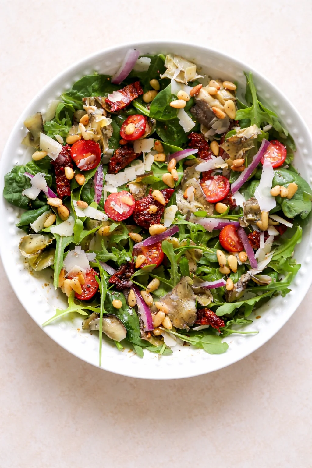 Bright arugula salad with cherry tomatoes, red onion, artichoke, pine nuts, and shaved parmesan in a white bowl.