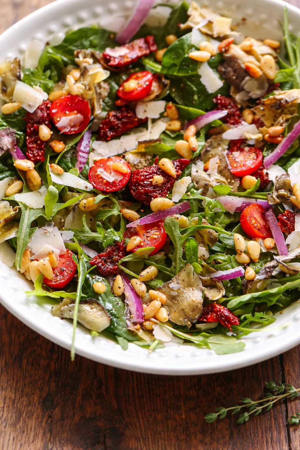 White bowl of arugula salad with cherry tomatoes, red onions, sun-dried tomatoes, parmesan shavings, and pine nuts