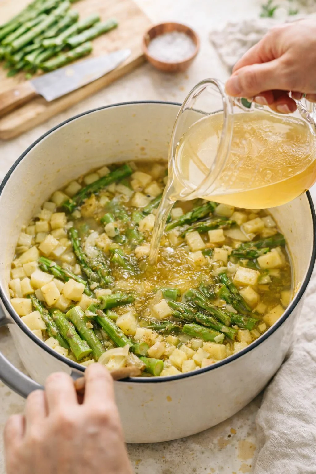 Hands pour broth into a cream-colored enamel pot with diced potatoes and green asparagus.