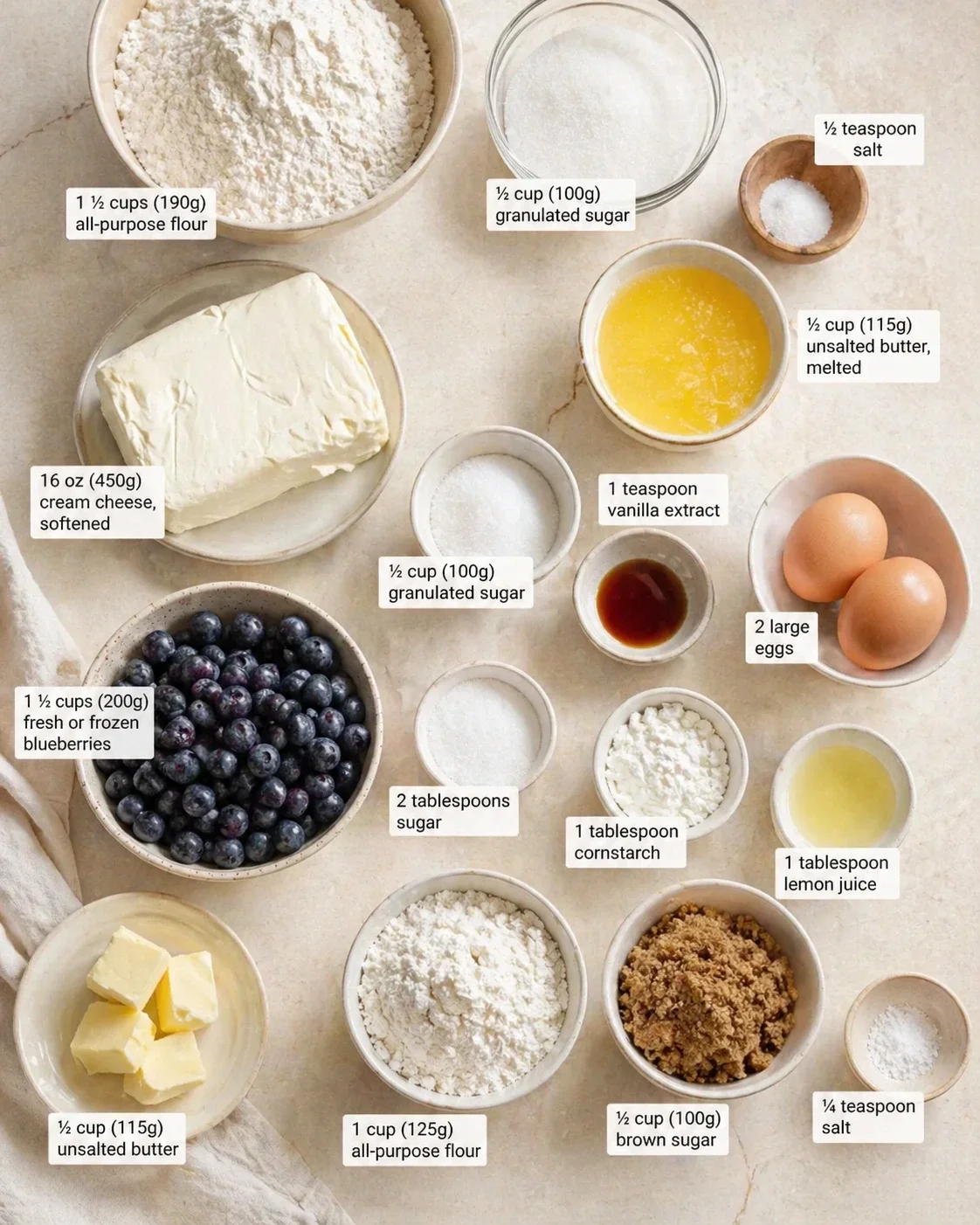 Overhead view of blueberry cheesecake ingredients in bowls and measuring cups arranged on a light marble surface