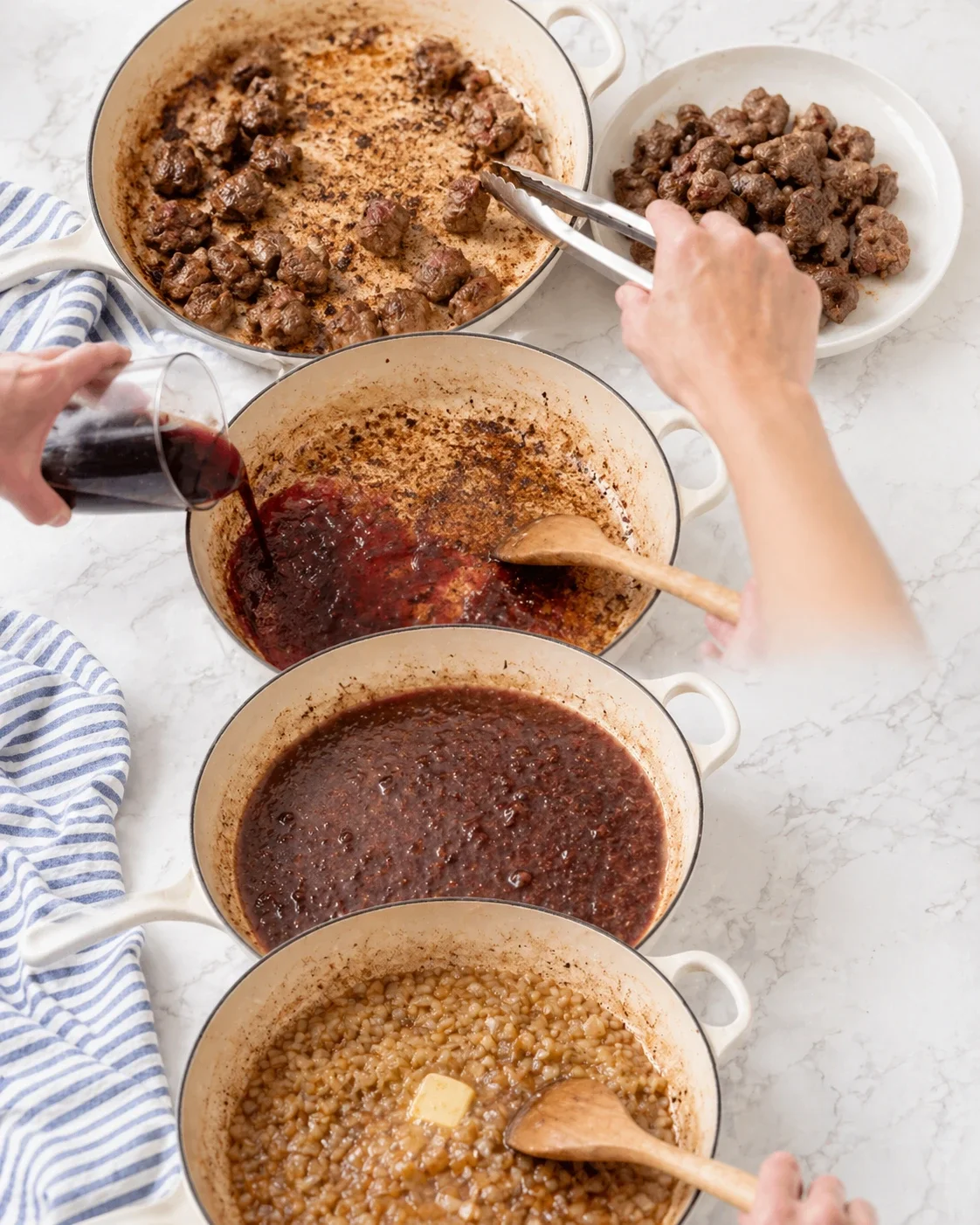 Overhead view of beef chunks browning in pots, with a hand pouring red wine into one pot.
