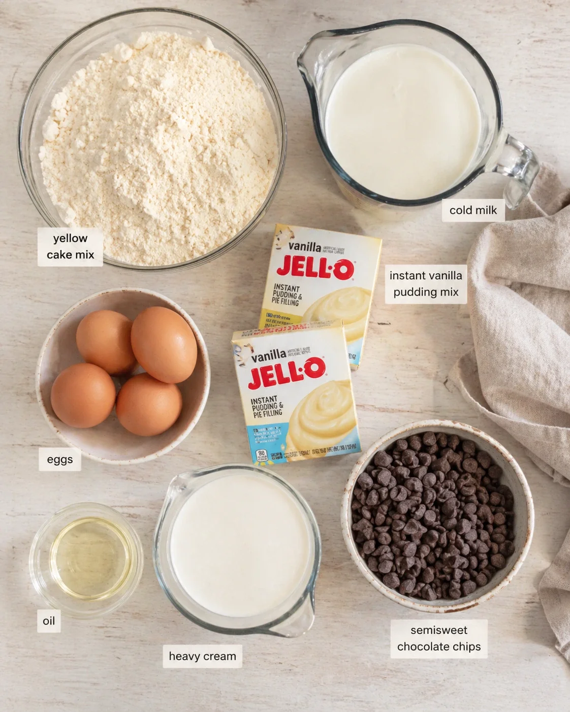 overhead view of cake-mix, pudding mix, milk, eggs, oil, cream, and chocolate chips laid out on a light wooden surface