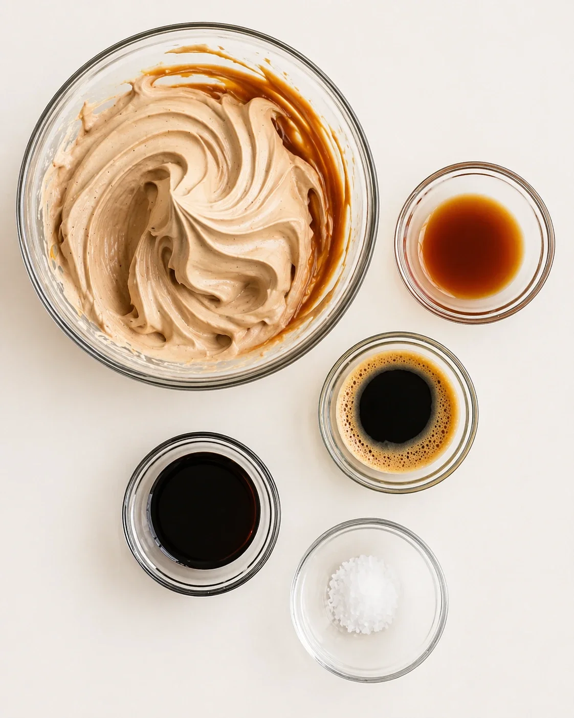 Overhead view of swirled beige frosting with caramel, coffee, and salt in small bowls around.