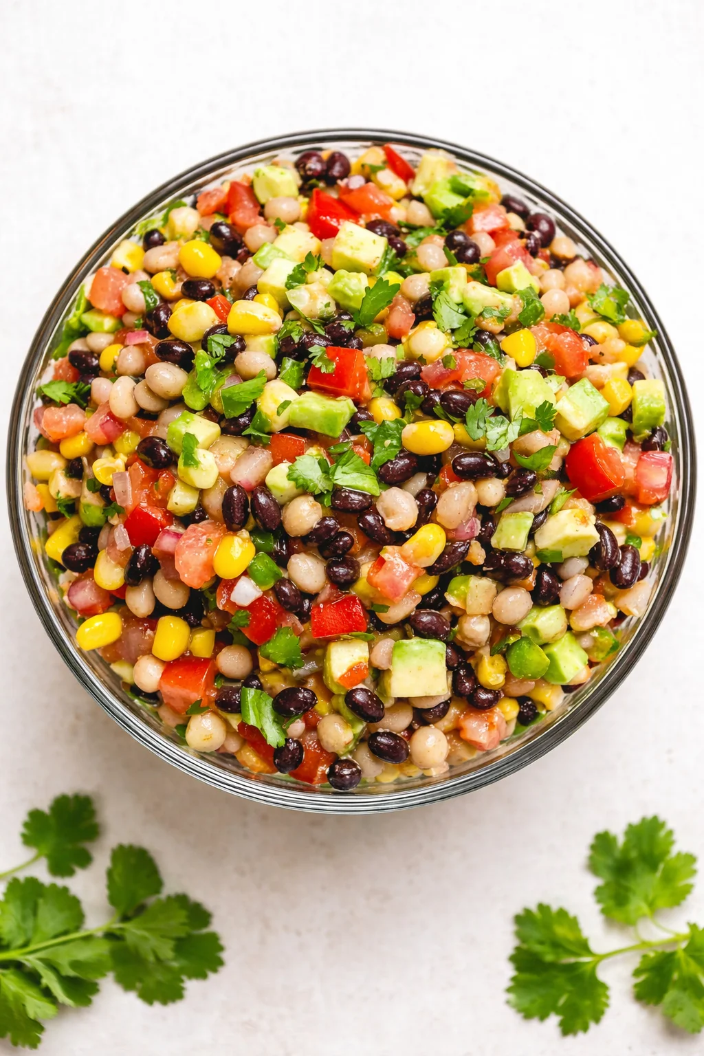 Top-down view of a glass bowl filled with colorful bean salad including avocado, corn, tomatoes, and cilantro.