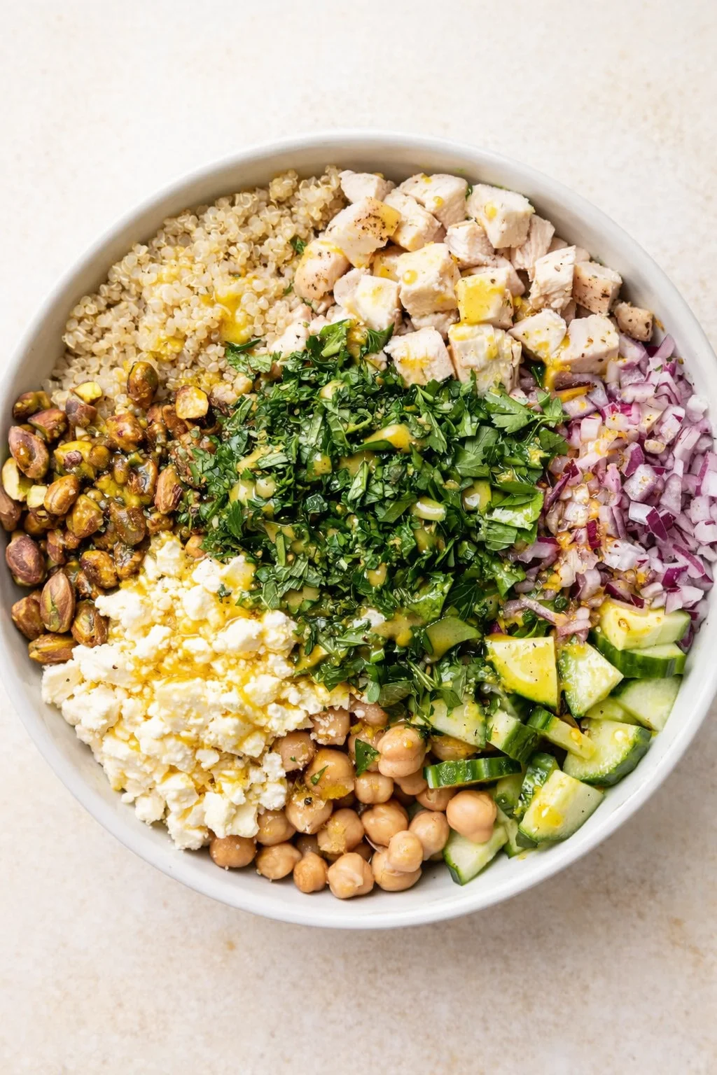 top-down view of a white bowl with quinoa, chickpeas, feta, diced chicken, avocado, cucumber, red onion, and herbs