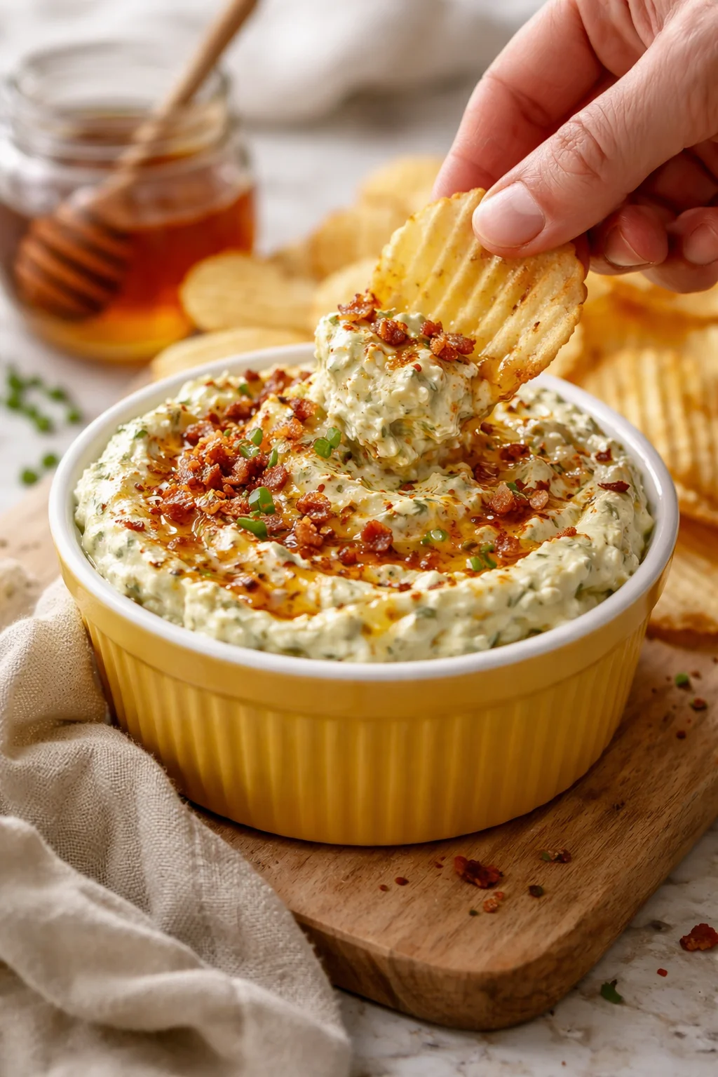 Golden-yellow ramekin of creamy spinach-artichoke dip topped with bacon bits and chives, a ridged chip dipped into it.