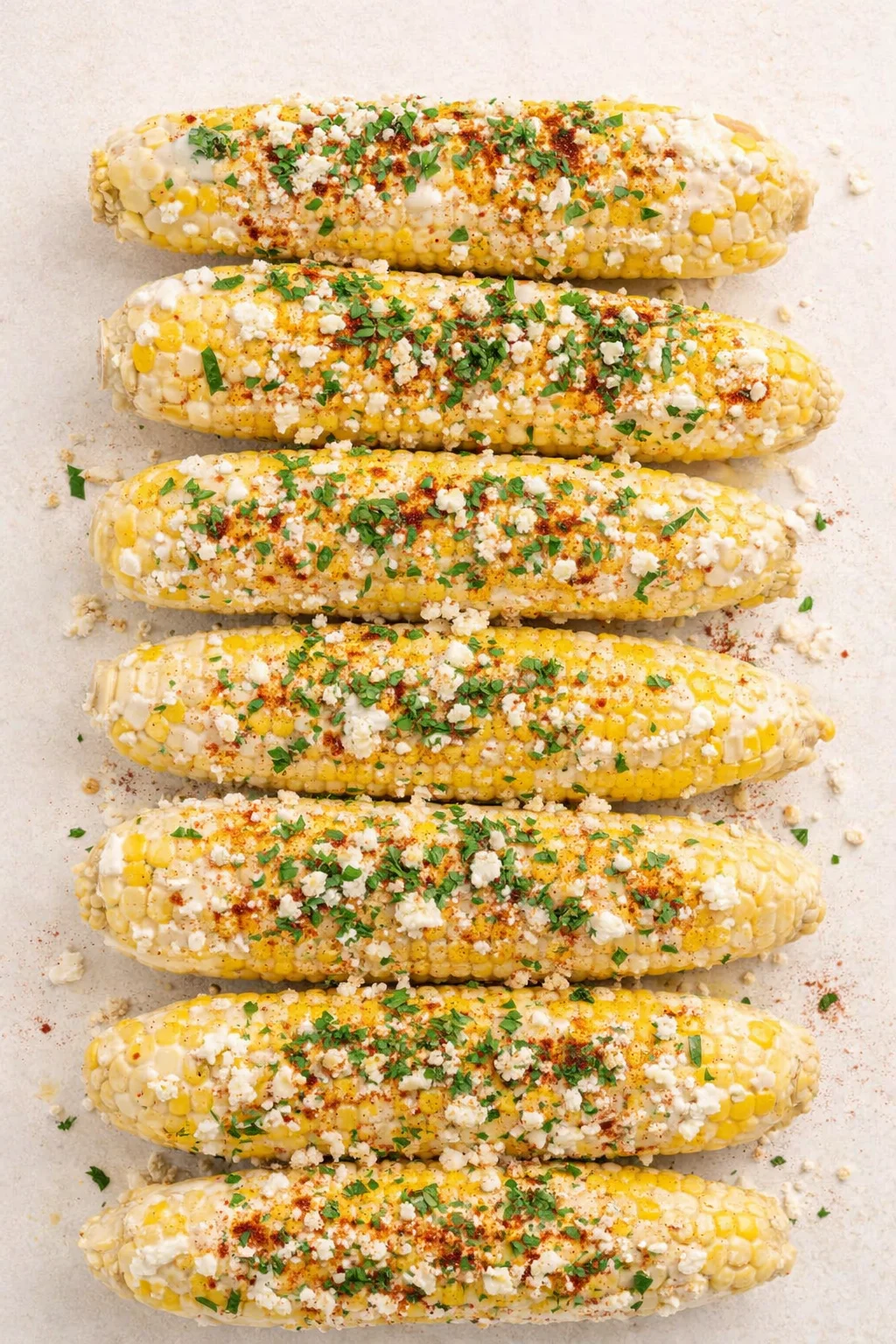 six ears of elote-style corn on a light surface, topped with crumbled cheese, cilantro, and spices