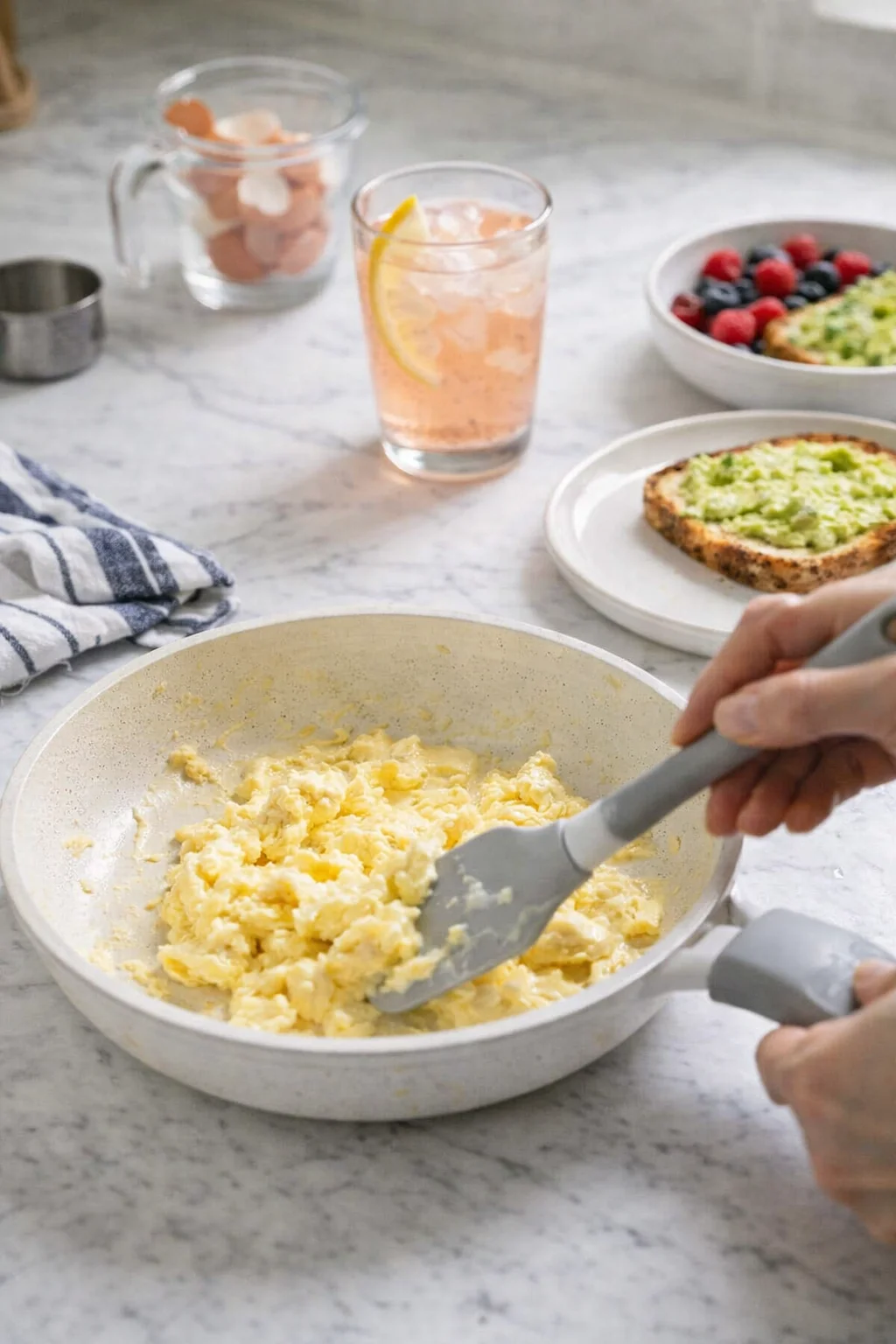 Creamy scrambled eggs in a white skillet on a marble counter with fruit toast and berries nearby.
