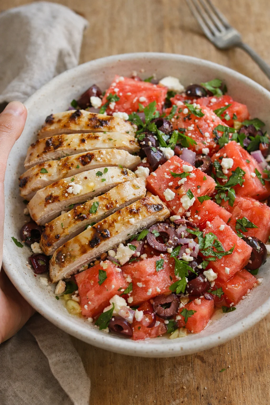 Sliced grilled chicken on a watermelon-feta-olive salad in a speckled ceramic bowl.