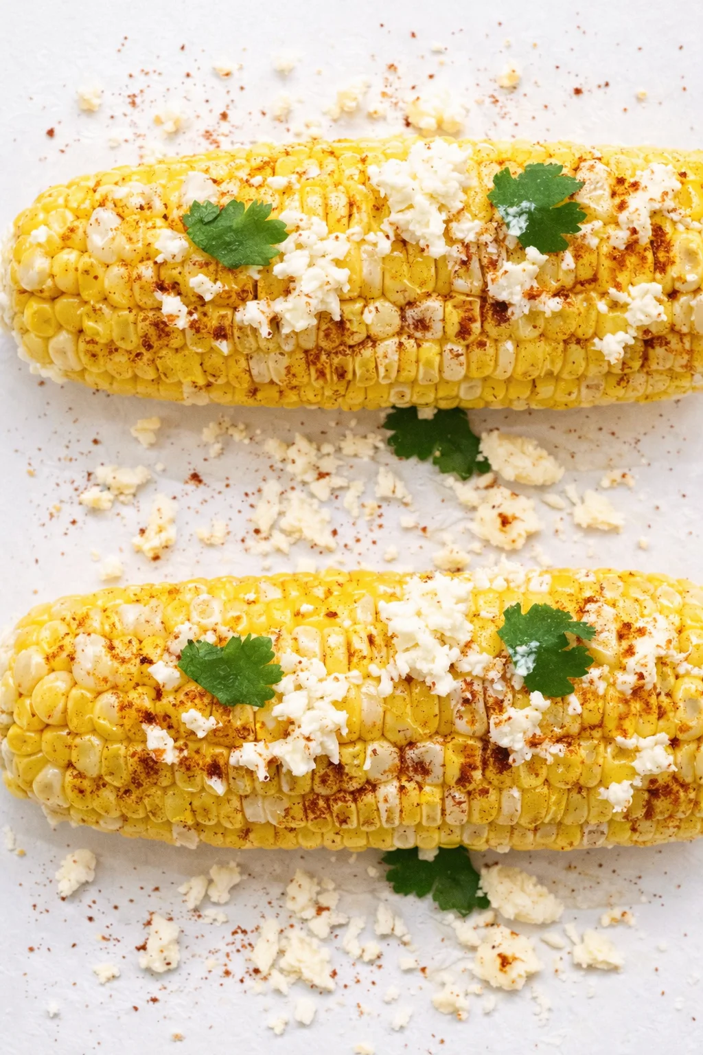 Two grilled corn cobs topped with crumbled cheese, chili powder, and parsley on a white surface.