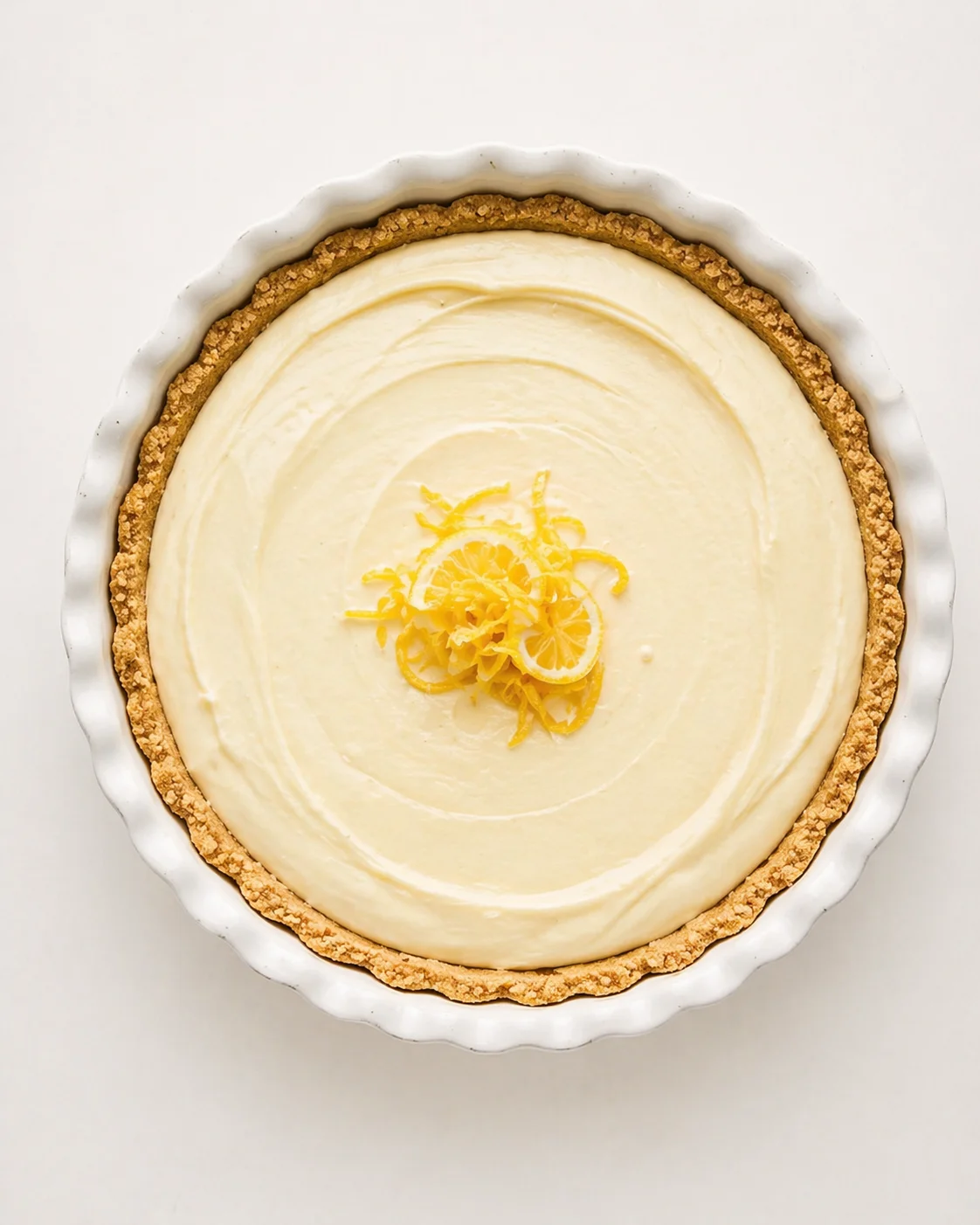 Round lemon cheesecake with crumb crust, topped with lemon zest in a white fluted dish.