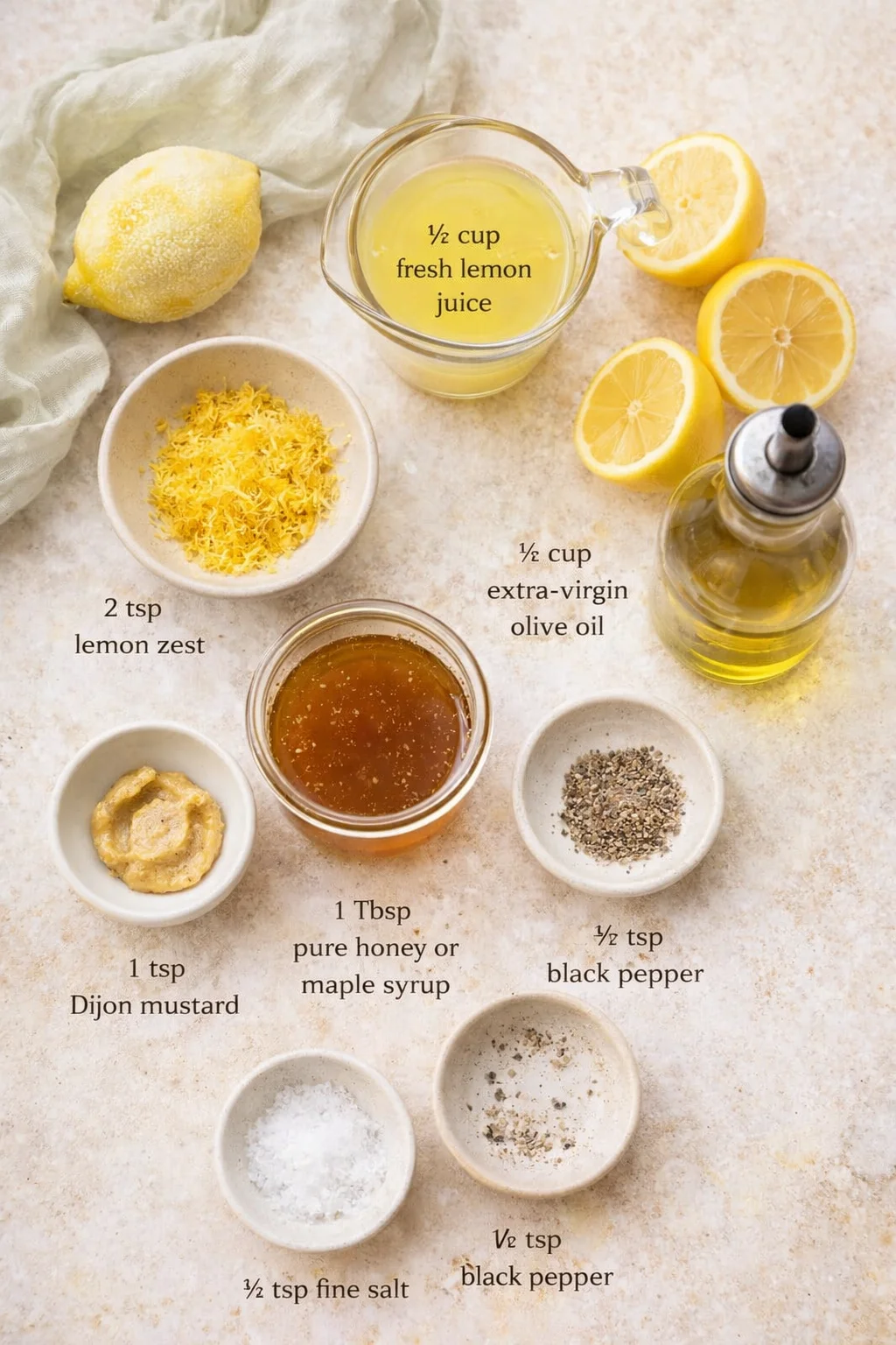 Overhead view of lemon juice, olive oil, lemon halves, zest, honey, mustard, salt, and pepper arranged on a light countertop.