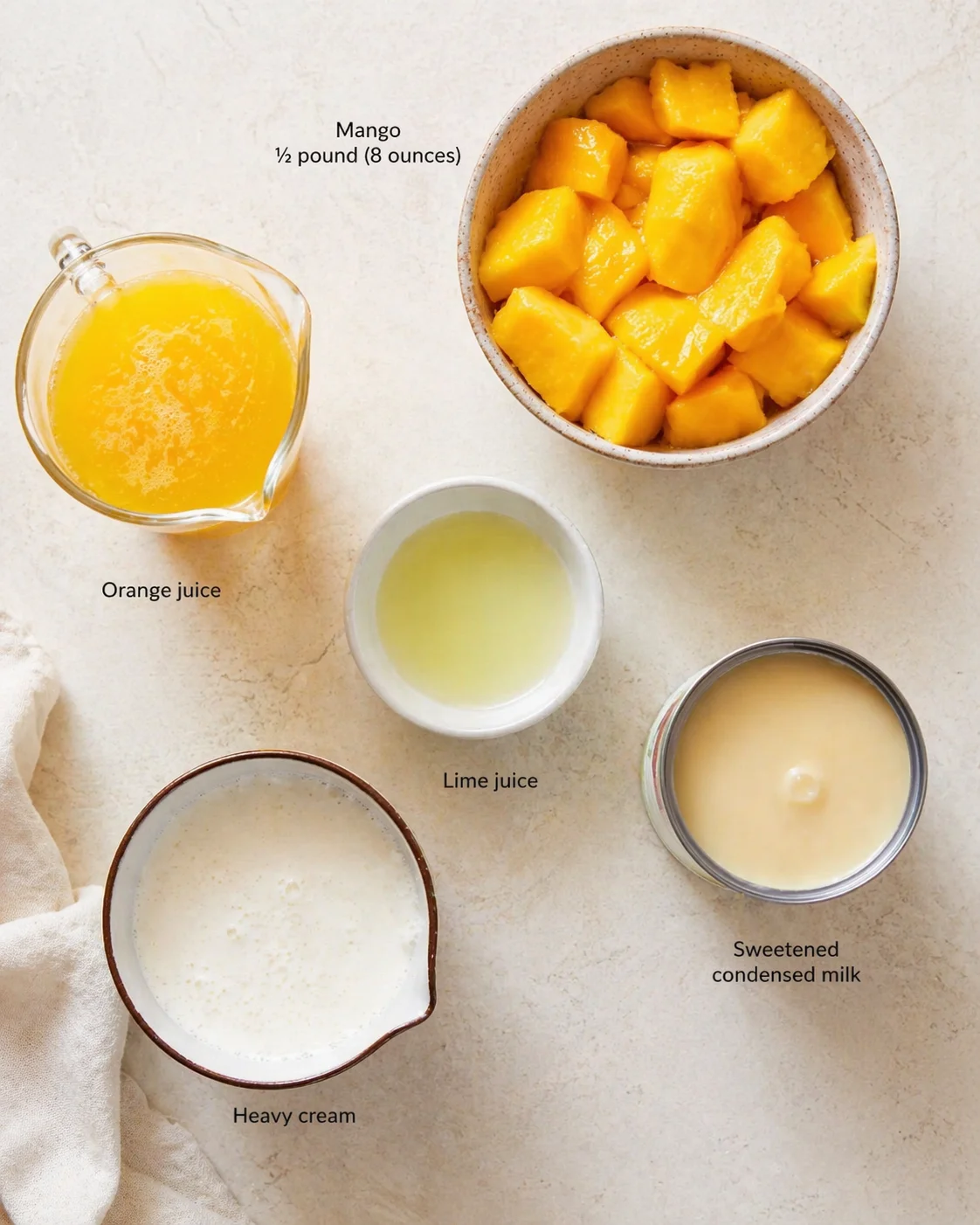 Overhead view of mango chunks, orange juice, lime juice, condensed milk, and cream on a beige surface