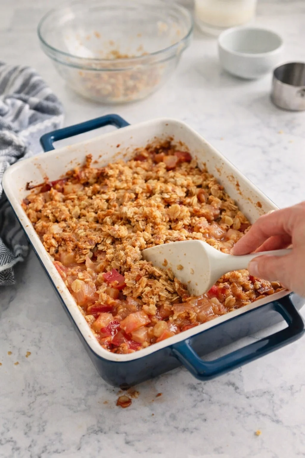 Blue enamel baking dish with golden oat-topped fruit crumble being scooped by a hand