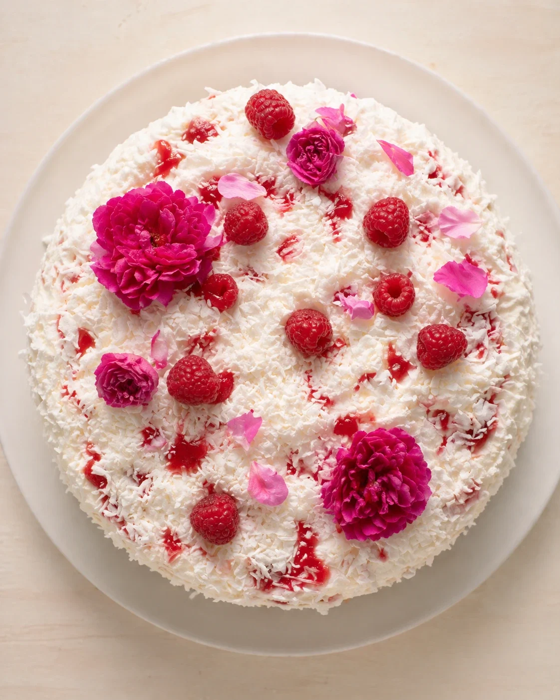 round cake coated in coconut with raspberries and pink flowers on a white plate