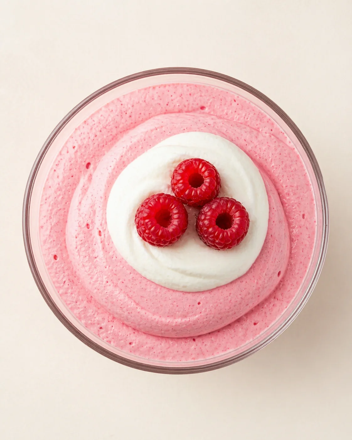 Top view of pink raspberry mousse with whipped cream and three raspberries in a glass bowl.