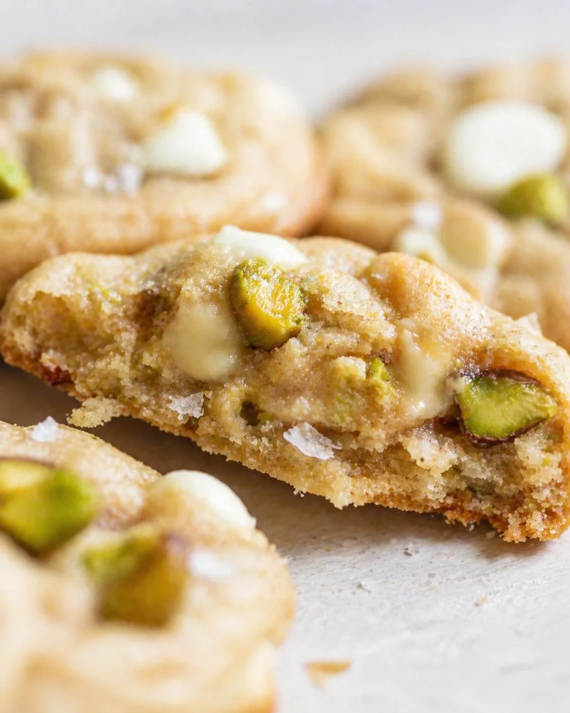 Close-up of pistachio-studded cookies with melted white chocolate on a light surface.