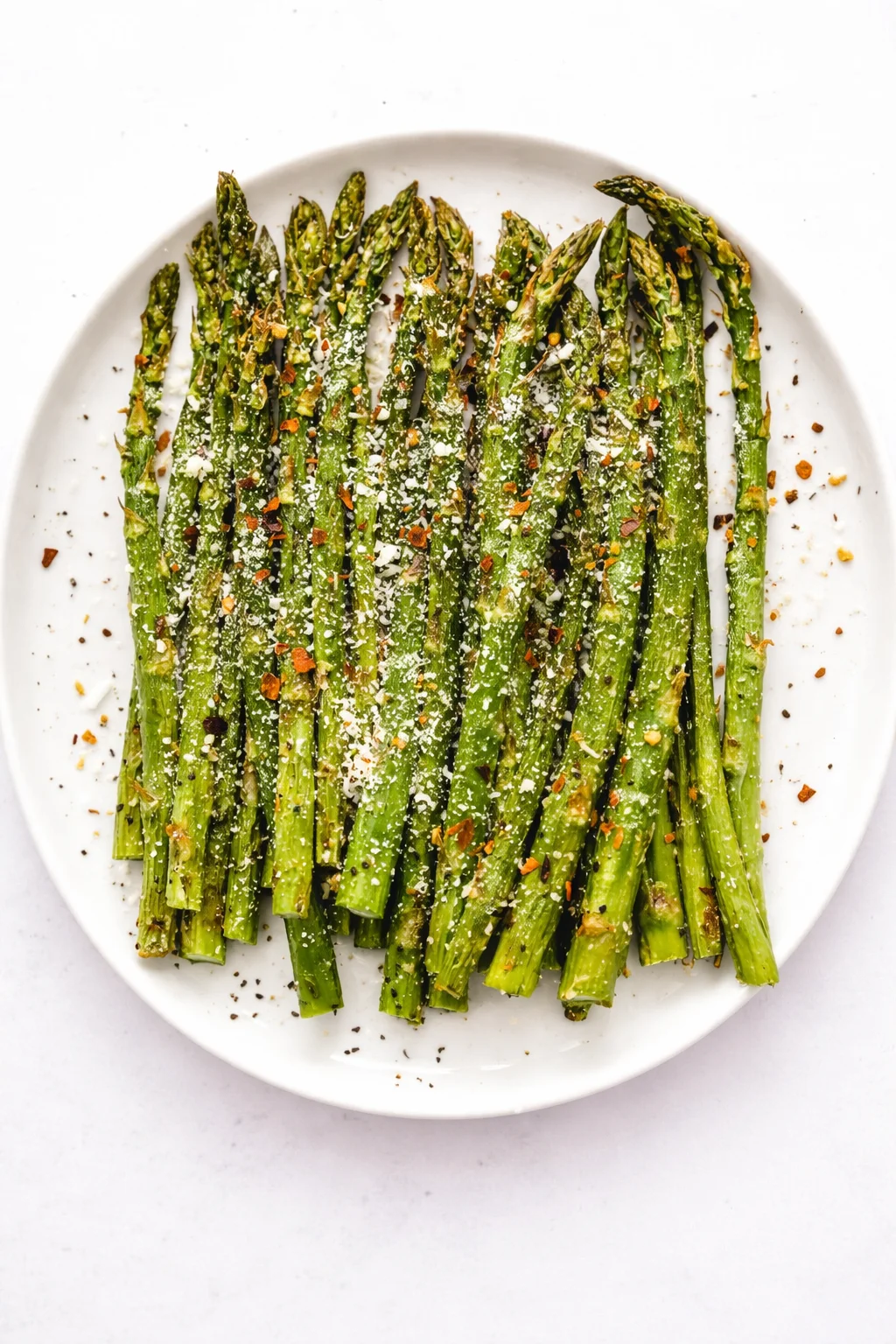 Bright green roasted asparagus spears on a white plate, topped with parmesan and red pepper flakes.