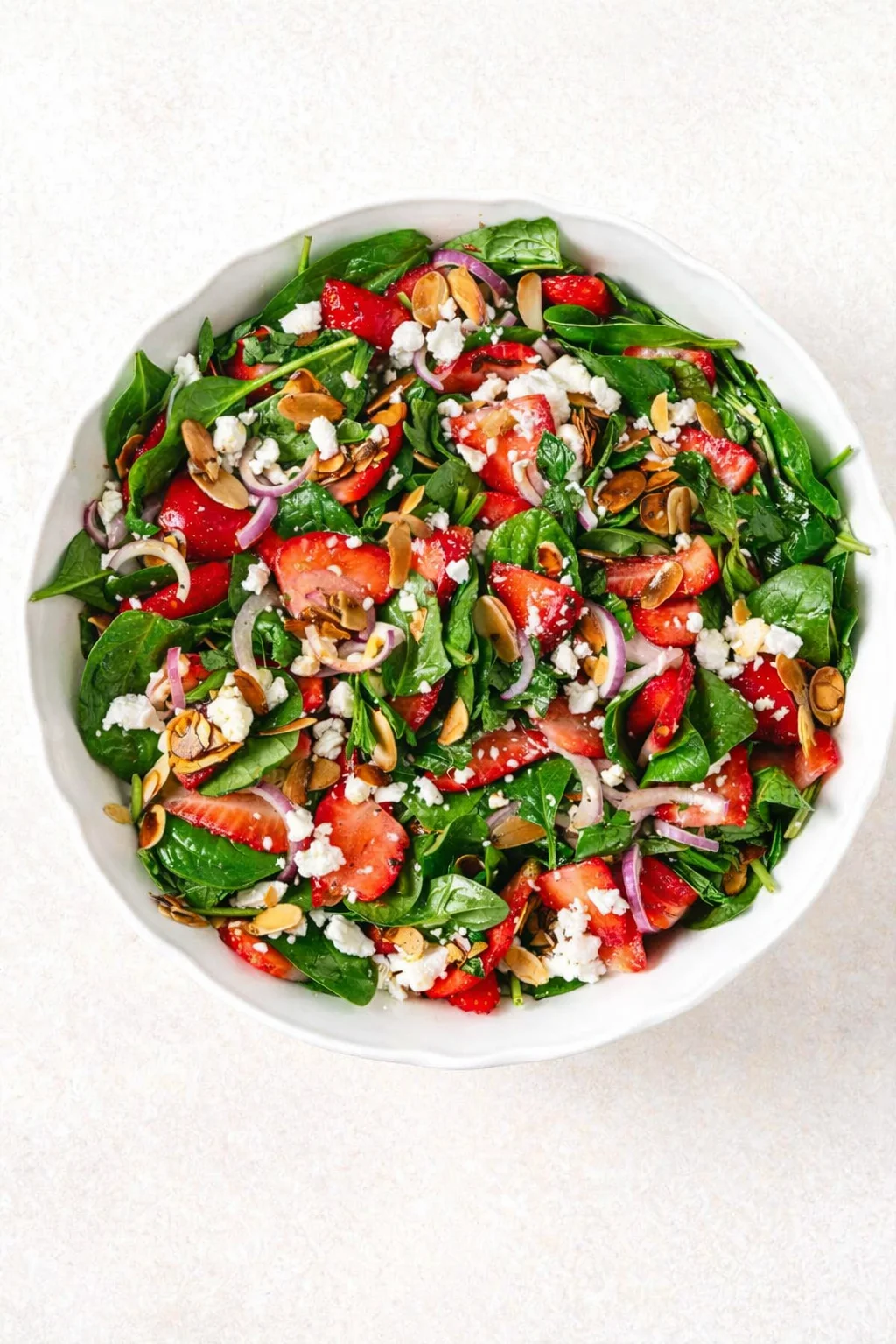 Round white bowl of spinach strawberry salad with feta, red onion, and toasted almonds on a light textured background.