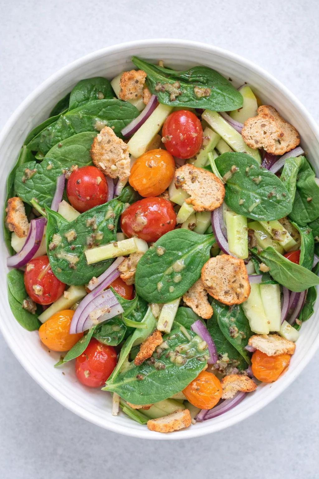 Top-down view of a spinach salad with cherry tomatoes, cucumber sticks, red onion, and croutons.