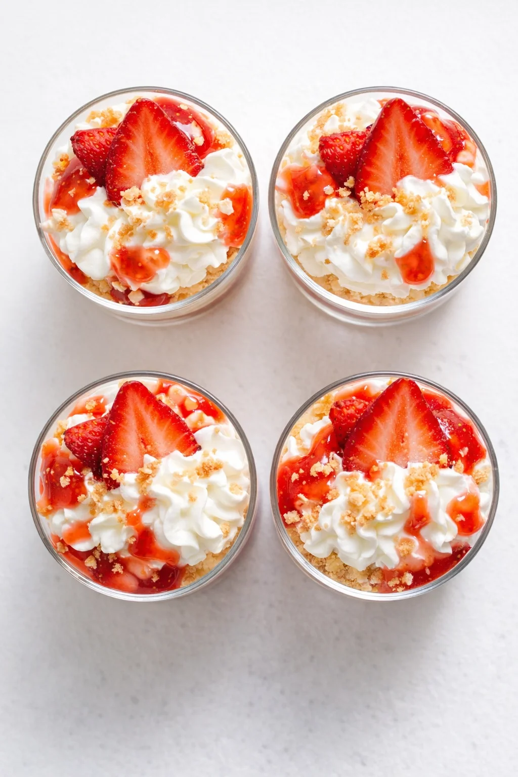 Four glass dessert cups topped with whipped cream, fresh strawberry slices, strawberry sauce, and crumb crust.
