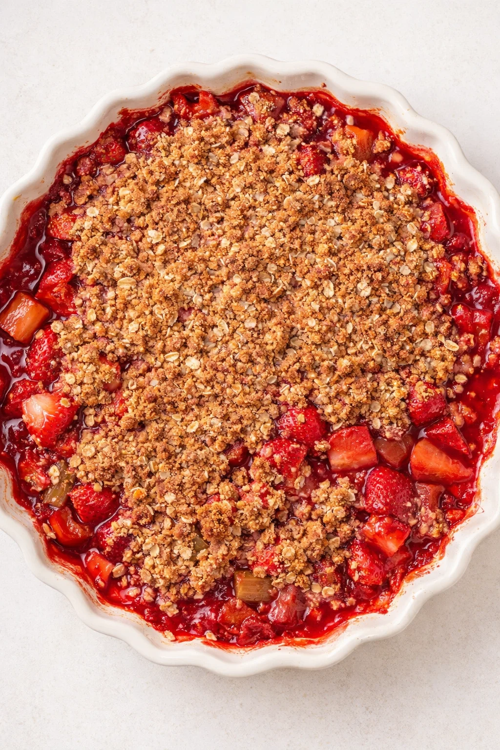 top-down view of a white fluted baking dish with red strawberry-rhubarb filling and golden-brown crumble topping