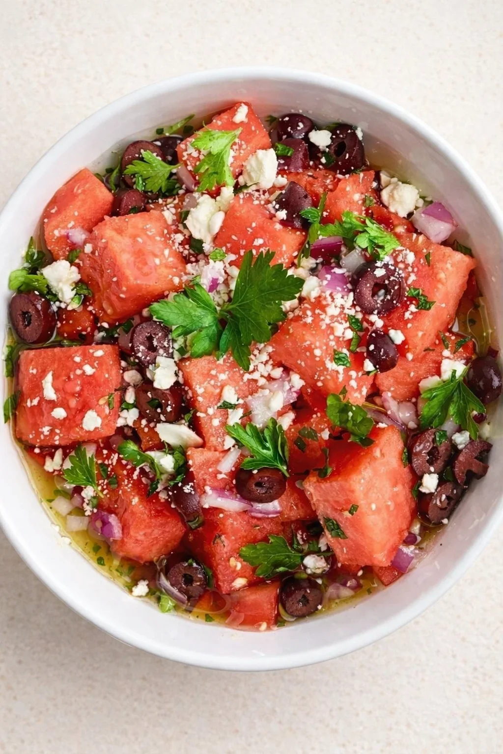 White bowl of watermelon feta salad with olives, red onion, and parsley on a light countertop.