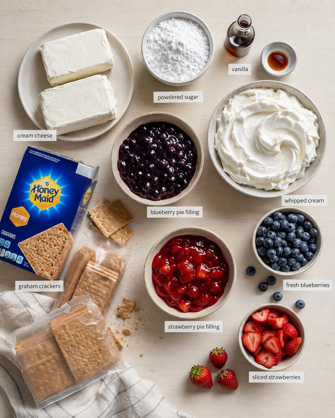 Overhead view of pie ingredients arranged on a wooden surface: cream cheese, powdered sugar, vanilla, whipped cream, berries, and crackers.