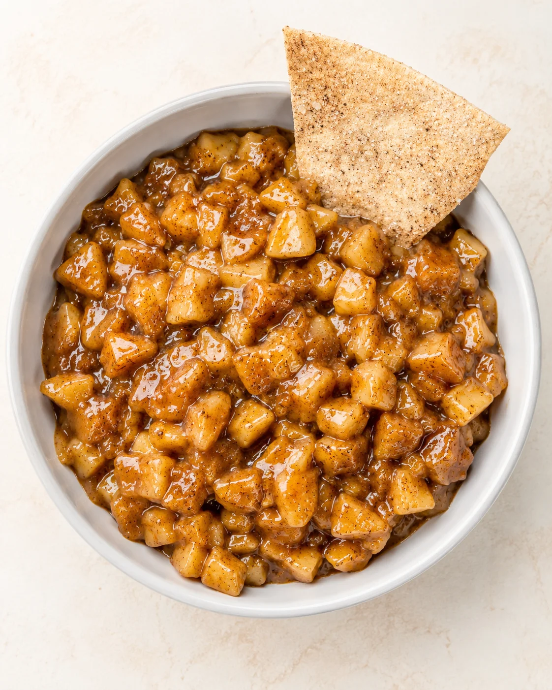 Top-down view of caramelized apple dice in glossy caramel sauce in a light-gray bowl with a crisp cracker