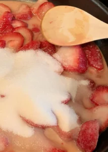 granulated sugar on top of cut fresh strawberries in a pan with a wooden spoon.