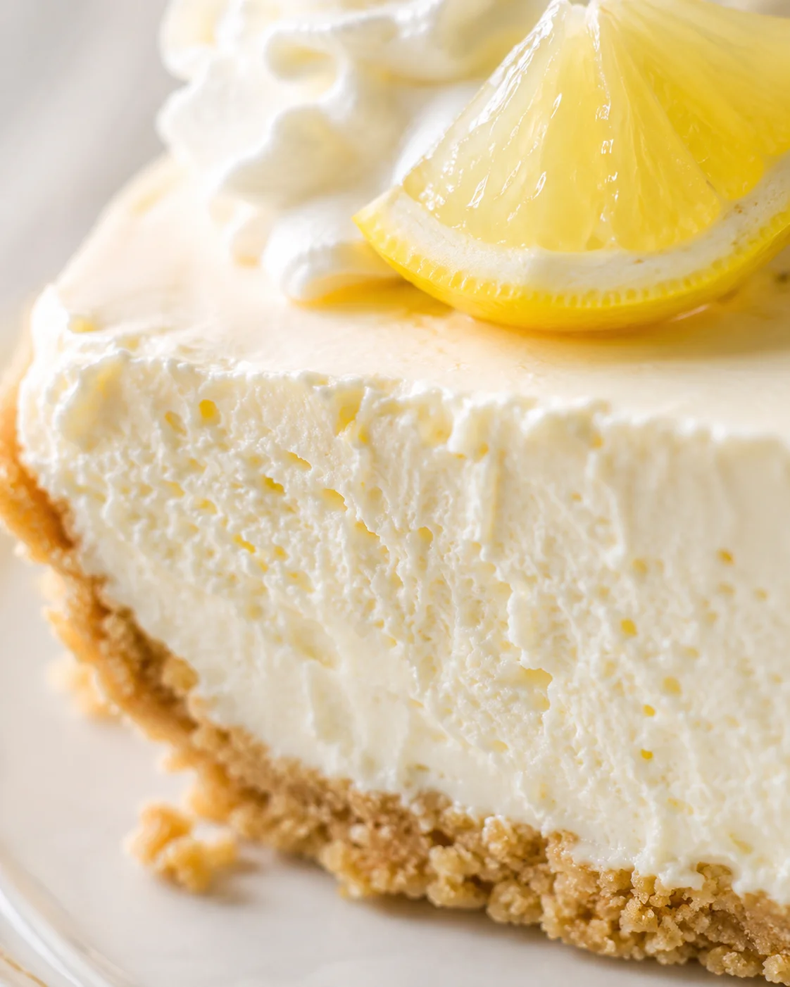 close-up of a lemon cheesecake slice with crumb crust, creamy filling, lemon wedge, and whipped cream