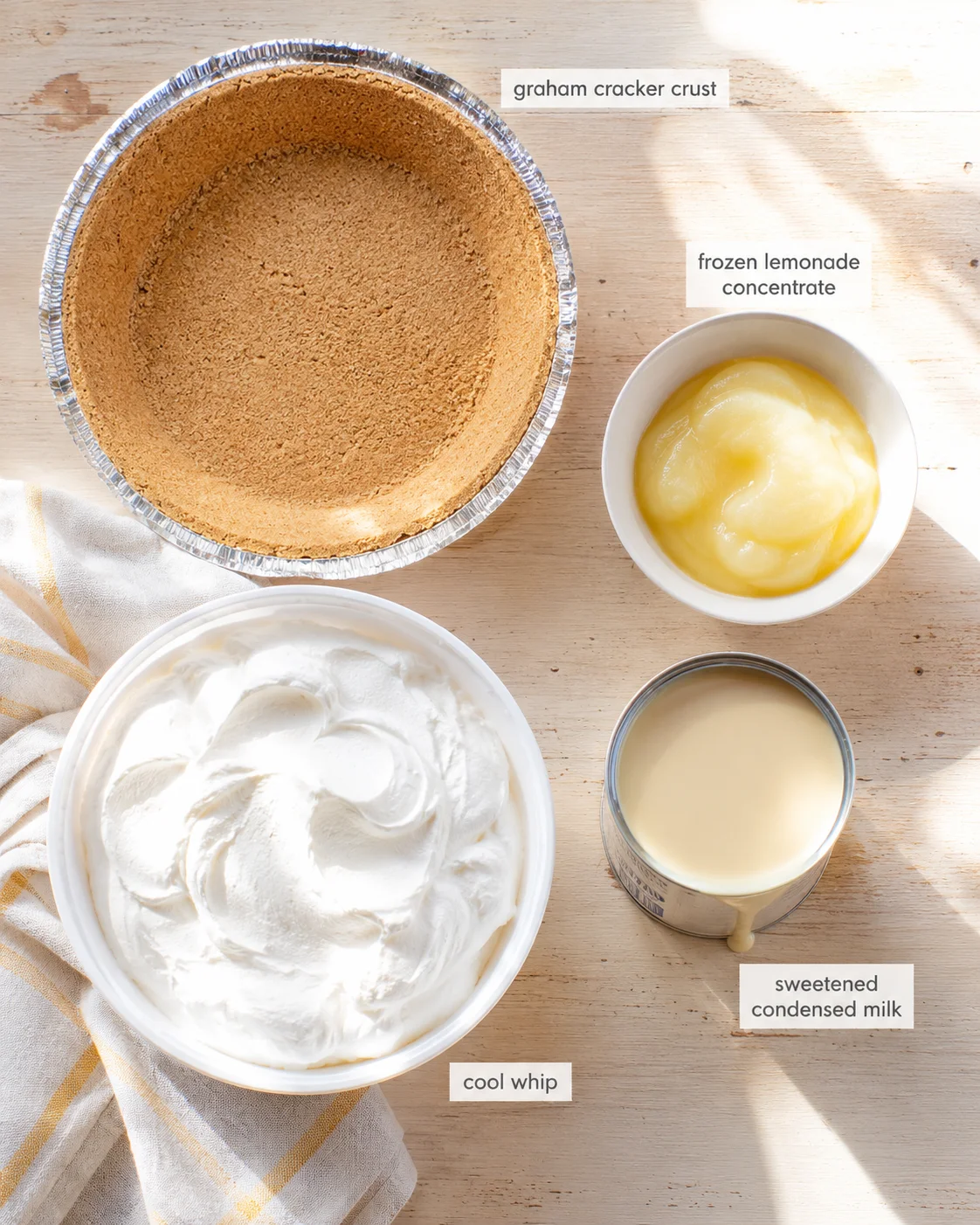 Top-down view of graham crust in a foil pan, whipped cream, lemon concentrate, and condensed milk on a wooden surface.