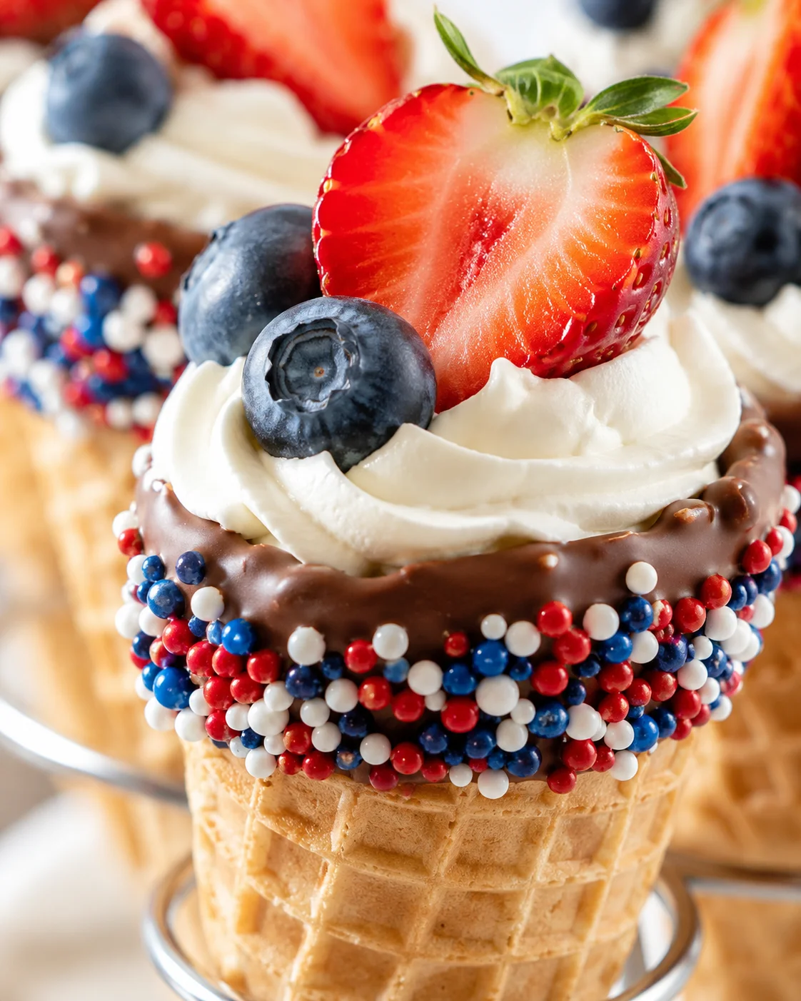 Close-up of a chocolate-dipped cone with whipped cream, strawberry slice, blueberries, and red-white-blue sprinkles.