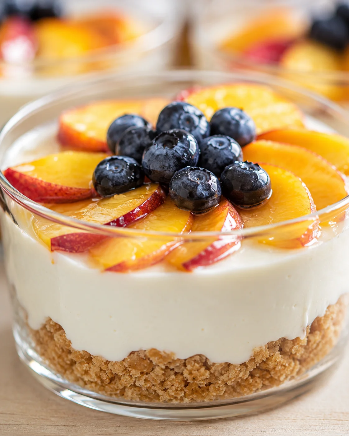 Creamy cheesecake with a crumb crust topped with peach slices and blueberries in a glass dish.